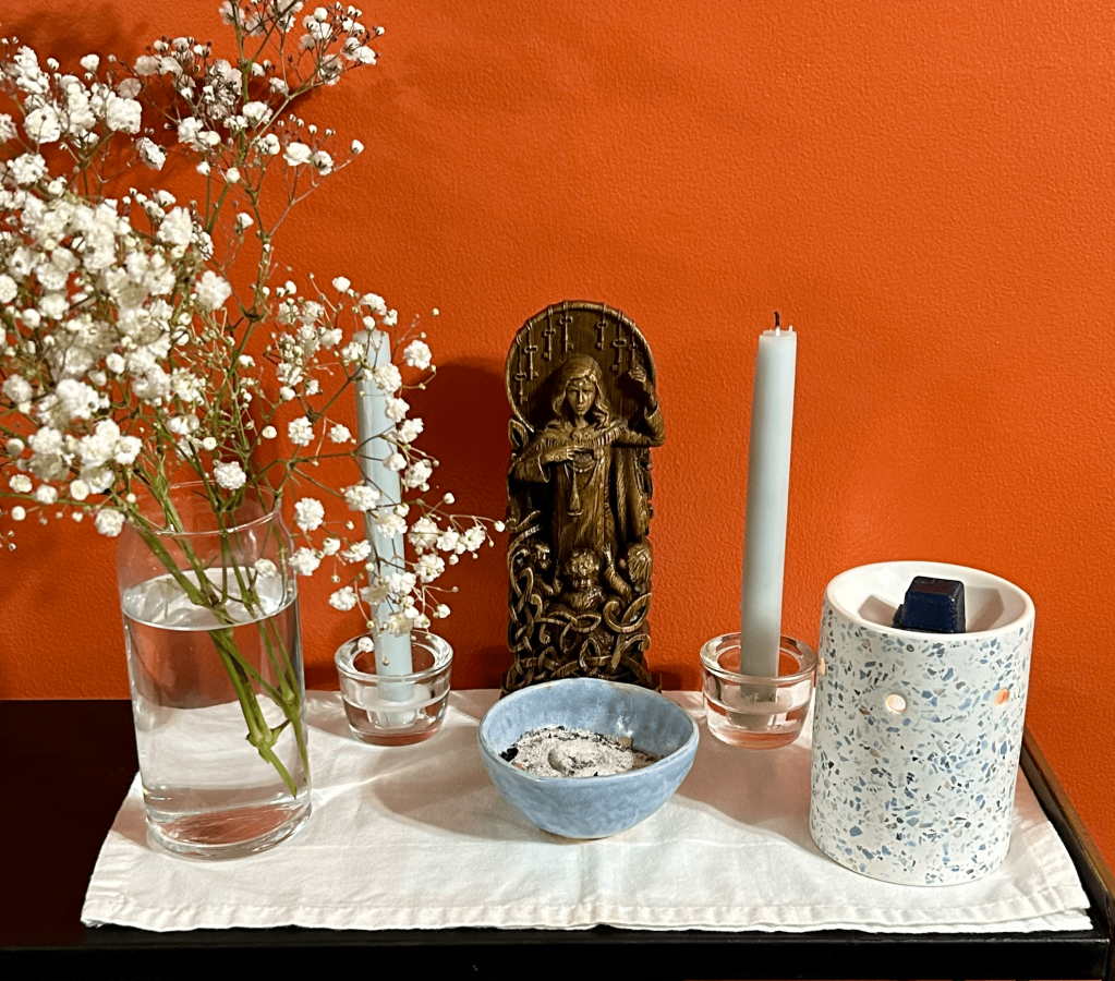 A statue of Frig using a drop-spindle and surrounded by children sits on a white cloth. Light blue candles flank the statue. A vase of baby's breath is to the left and a wax warmer to the right. An incense bowl filled with sand and ashes sits in front of the statue.