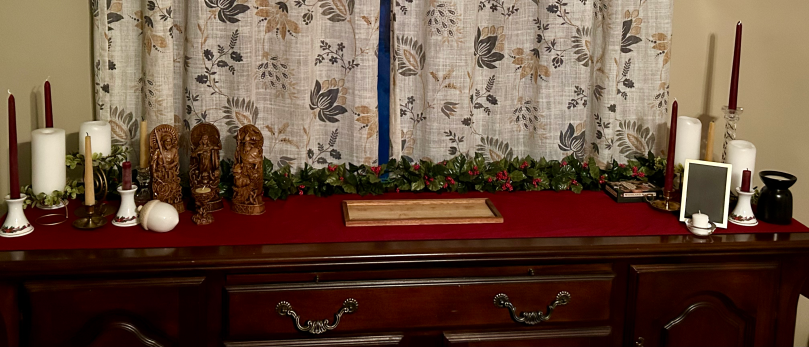 A sideboard with a deep red table runner. Each end has red, white and natural beeswax candles. On the left are statues of Ing, Frig, and Woden. On the right is a picture frame with black paper in it and a wax warmer.
