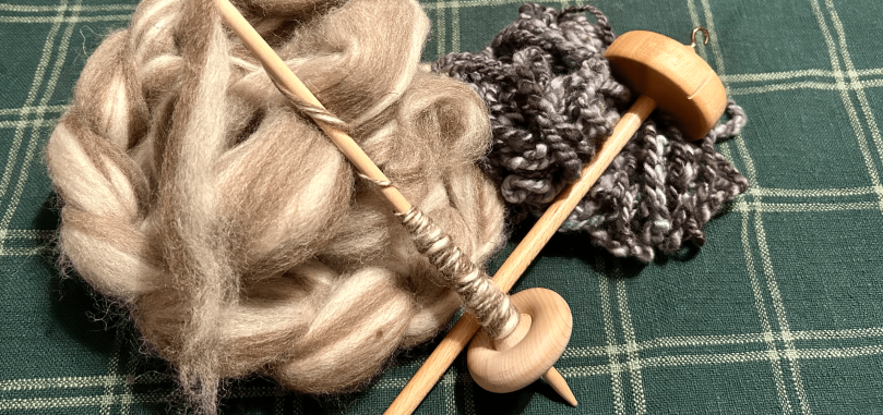 A top-whorl drop spindle and a bottom-whorl drop spindle crossed and resting against brown and cream wool roving and blue-gray hand-spun yarn.